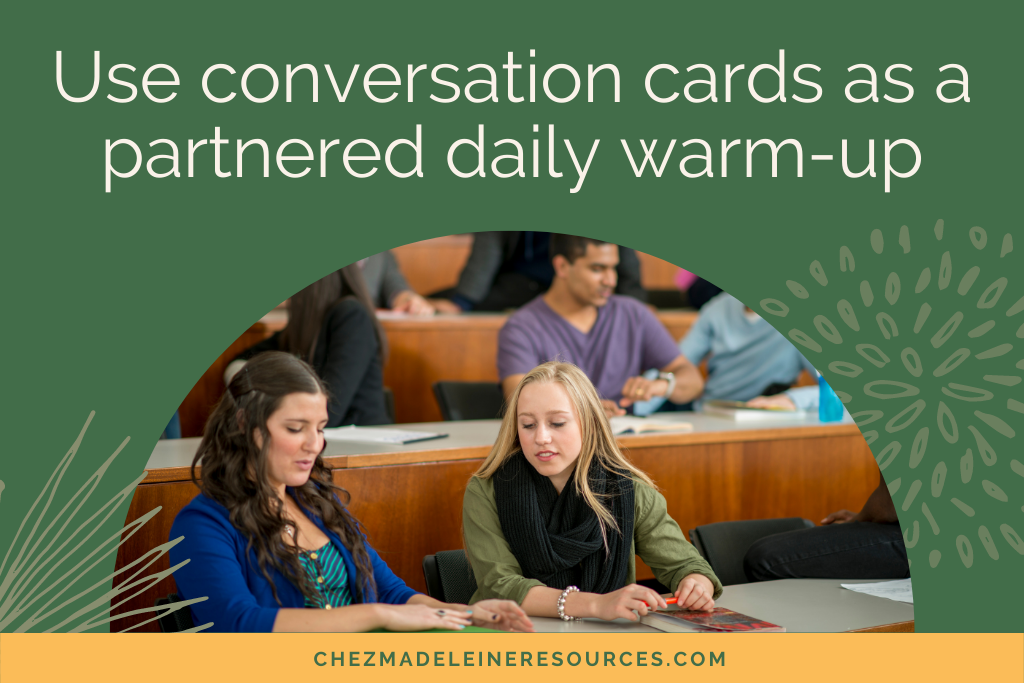 Two girls sitting in a classroom using conversation cards to practice their French. The text reads "use conversation cards as a partnered daily warm-up."